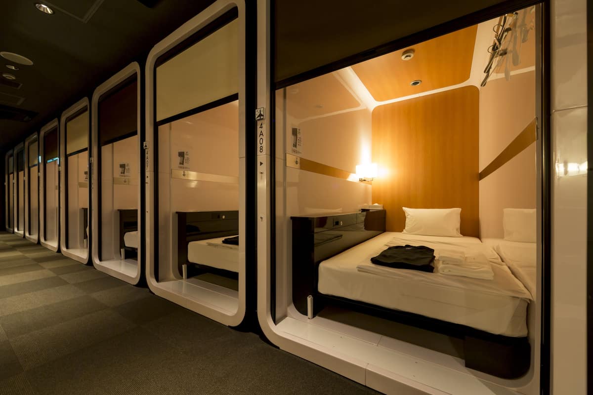 A row of compact, modern hotel rooms with individual beds, each featuring a small bedside table and a lamp.