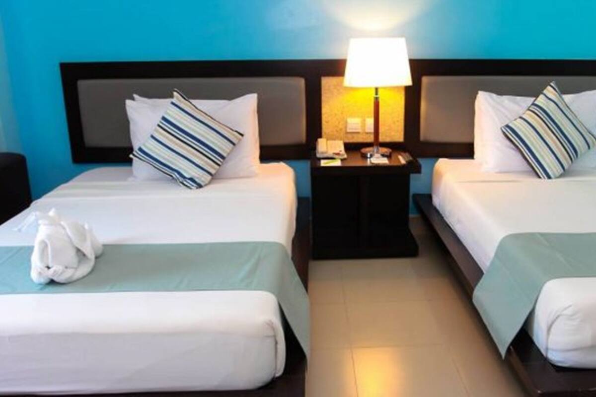 A hotel room with two beds, each with a striped pillow, a bedside table with a lamp, and a towel on the bed.