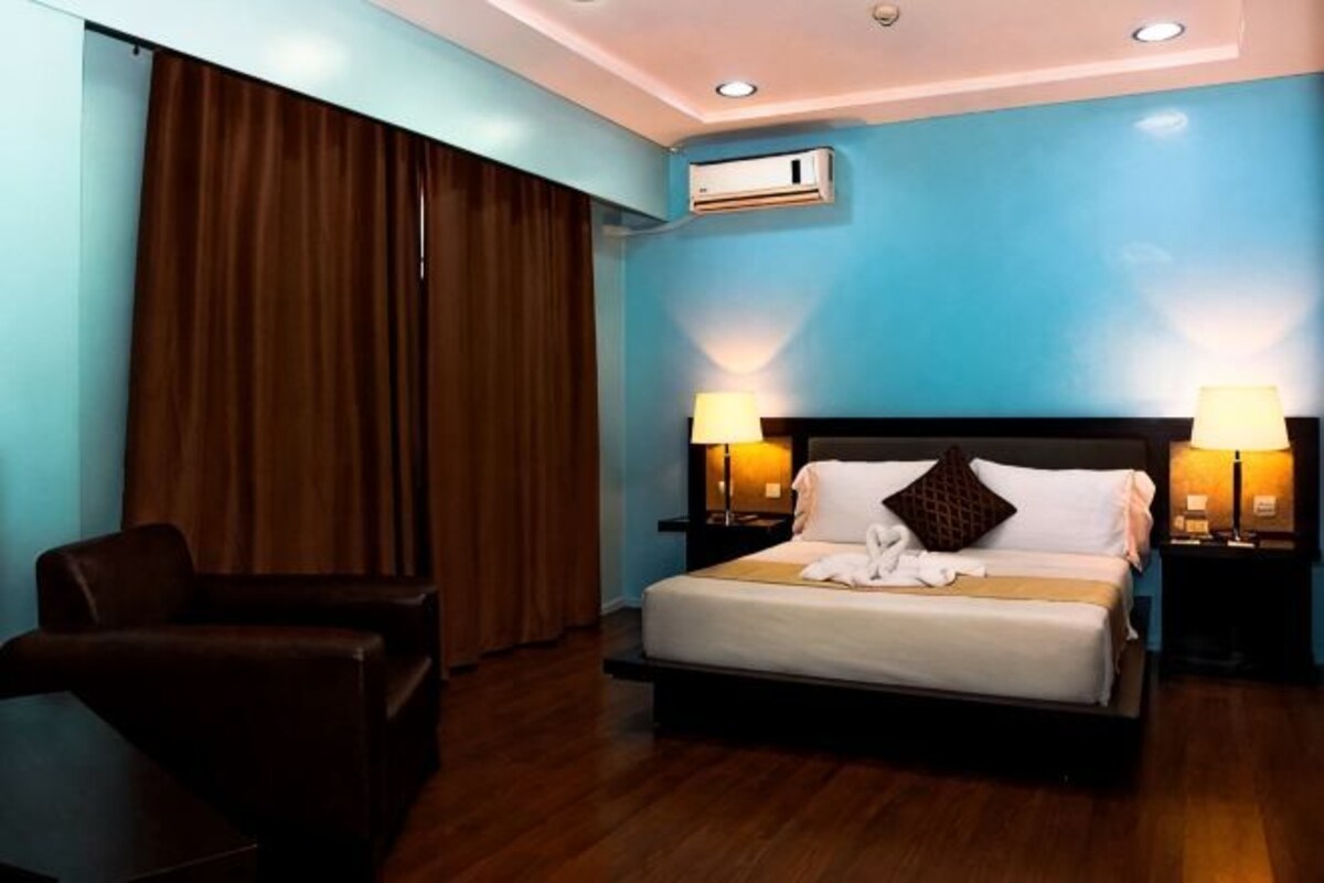 A hotel room with a bed, two bedside lamps, a chair, a window with curtains, and an air conditioning unit.
