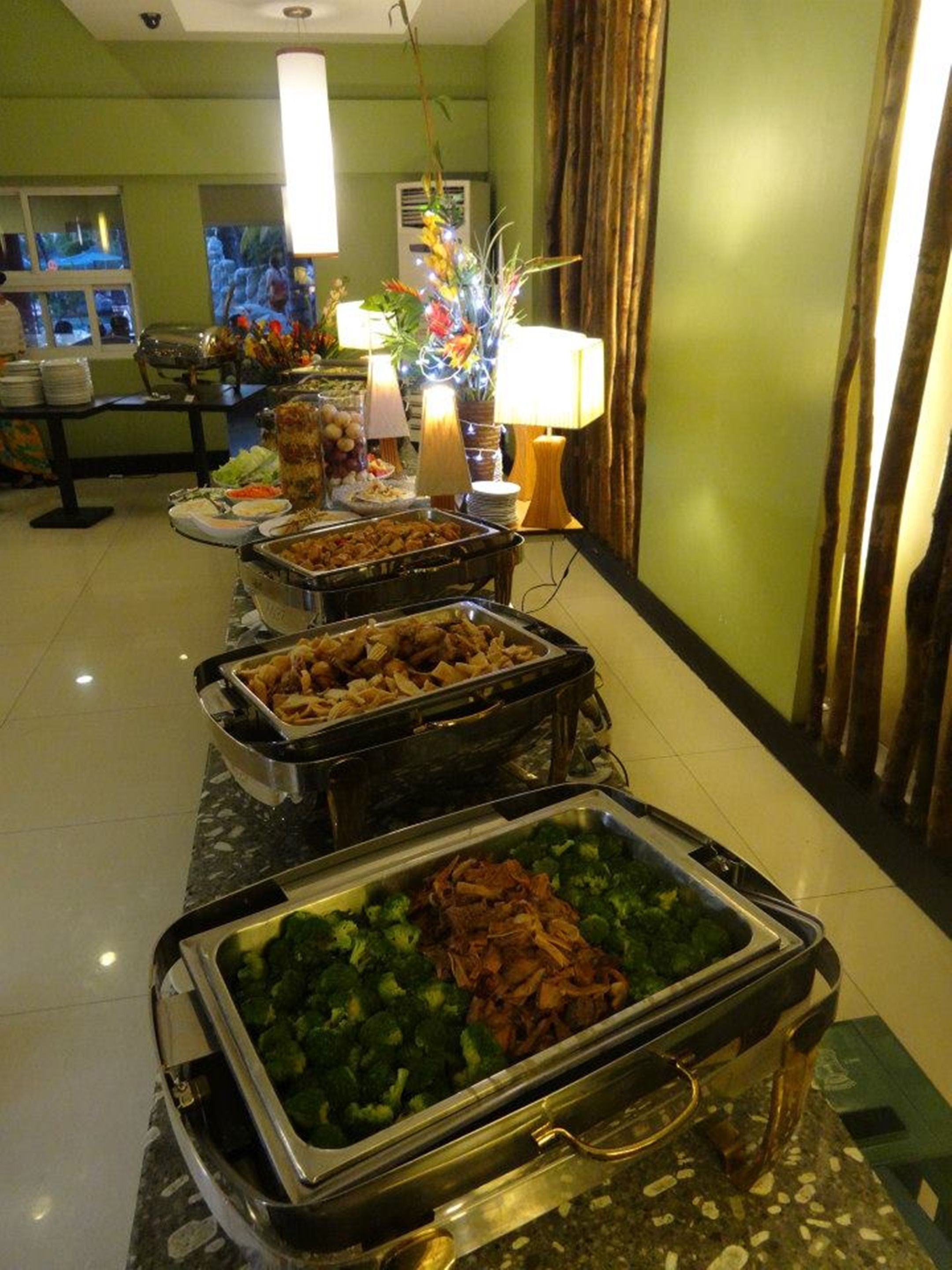 A buffet setup with various food items in metal chafing dishes, including vegetables and meats, in a hotel room with green walls and a tiled floor.
