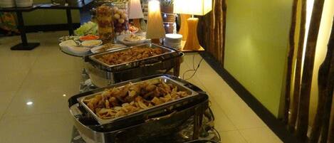 A buffet setup with various food items in metal chafing dishes, including vegetables and meats, in a hotel room with green walls and a tiled floor.