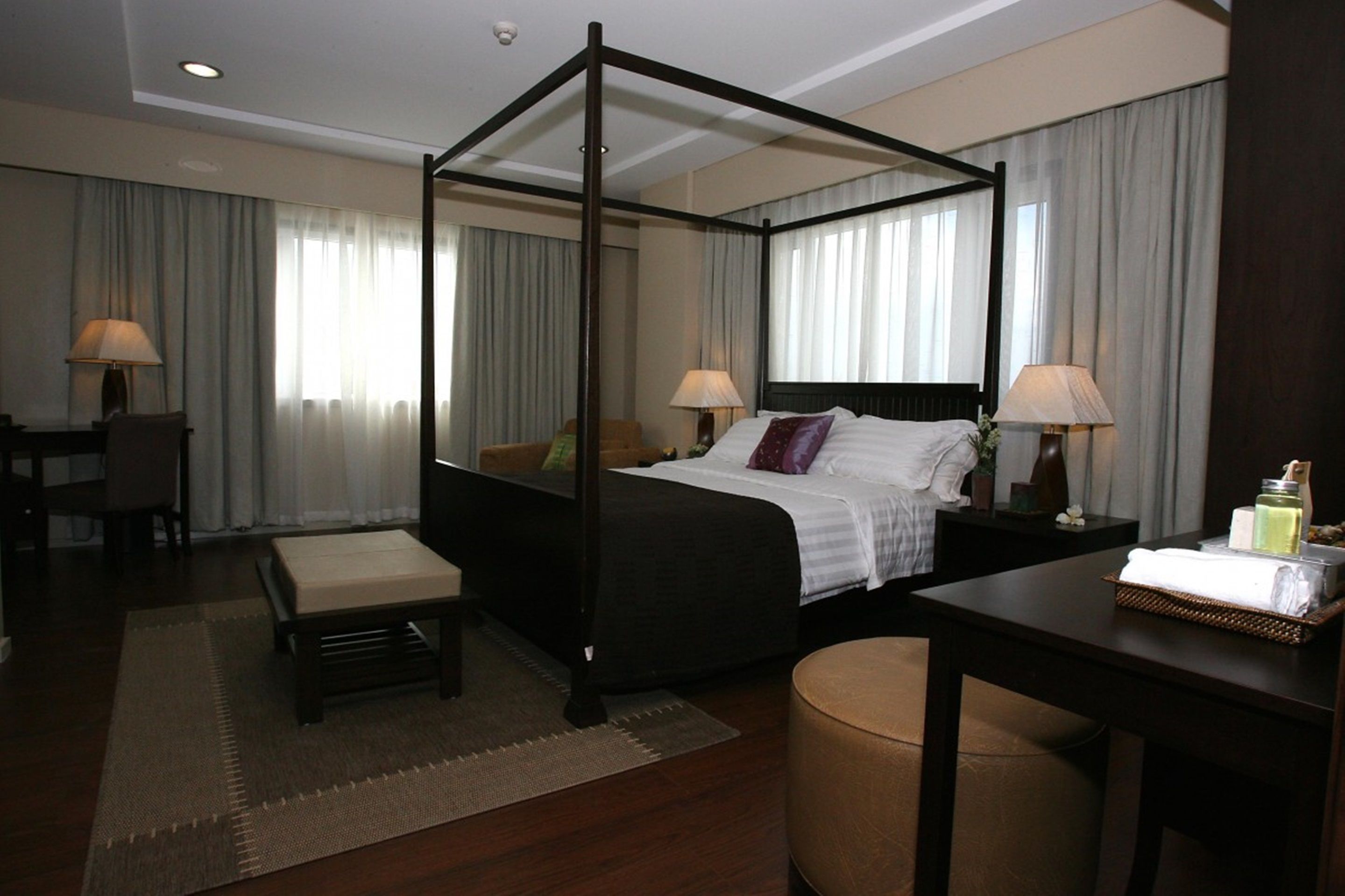 A four-poster bed with a dark frame, a bedside table with a lamp, a chair, and a small table with toiletries.