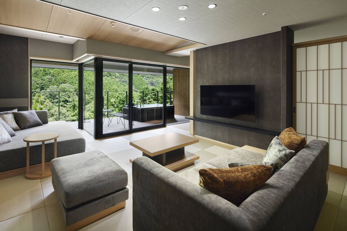 A modern living room with a sofa, ottoman, and coffee table, a flat-screen TV mounted on the wall, and a sliding glass door leading to a balcony with a view of greenery.