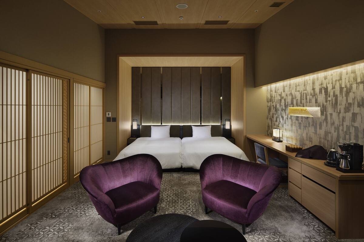 A hotel room with a large bed, two purple armchairs, a desk with a lamp, and a coffee maker.