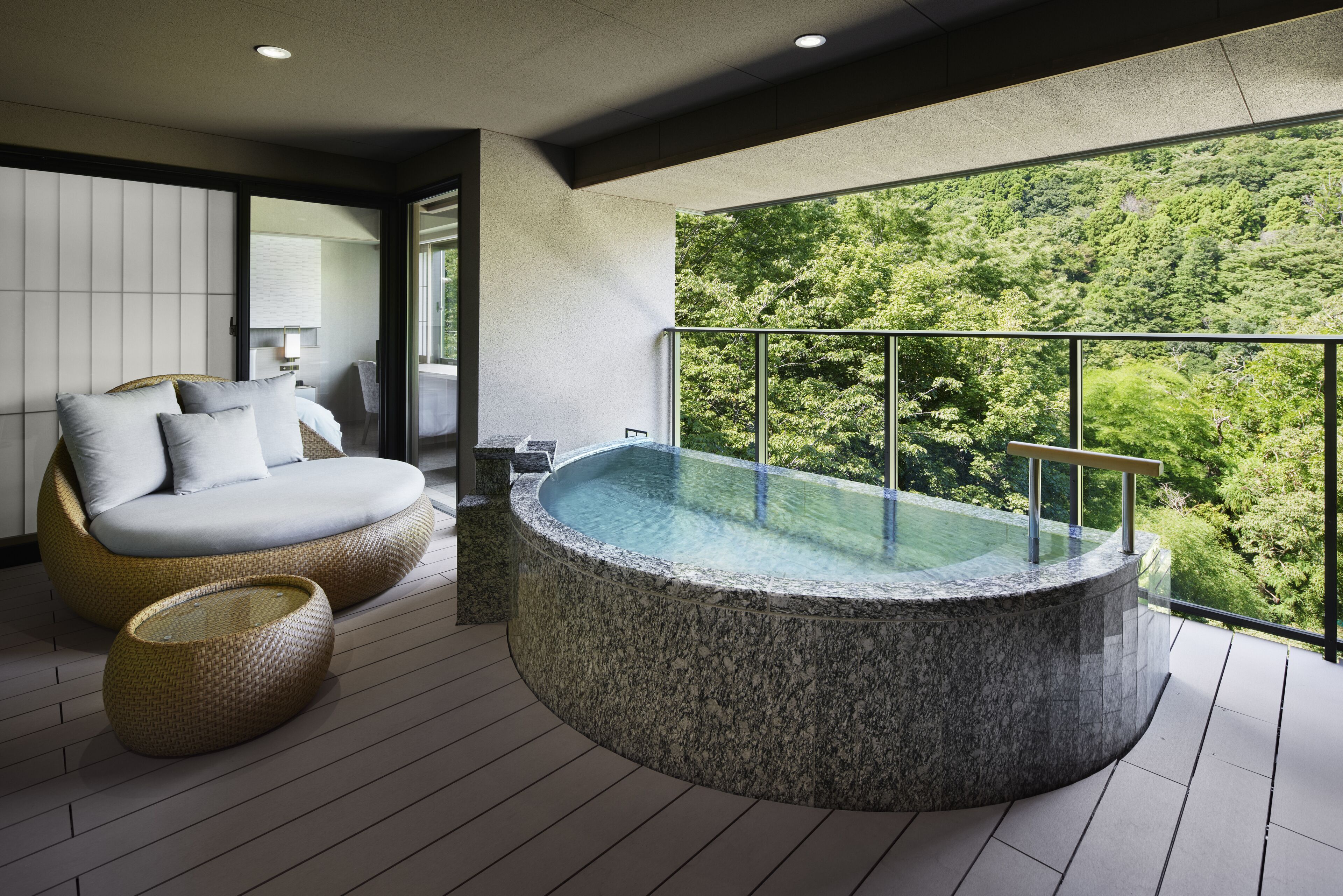 A modern outdoor bathroom with a hot tub, a cushioned seating area, and a view of a forested hillside.