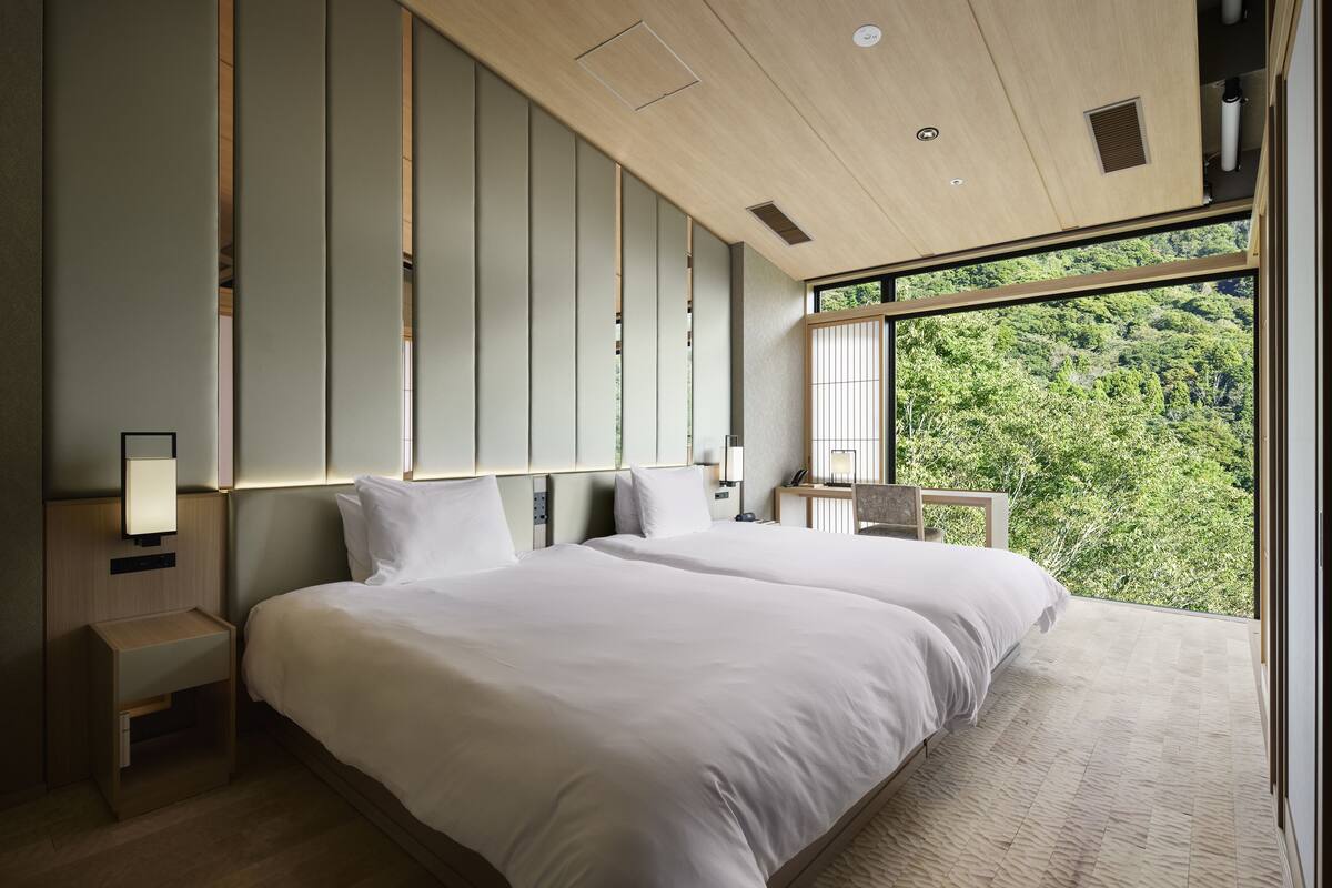 A modern hotel room with a large bed, a nightstand, a desk, and a view of a forest through large windows.