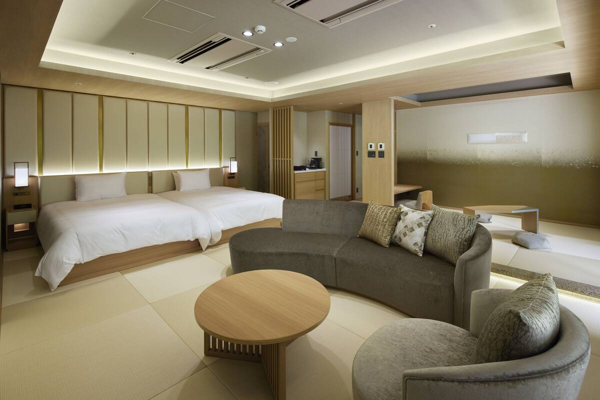 A modern hotel room with a large bed, a sofa, a round coffee table, and a small armchair.