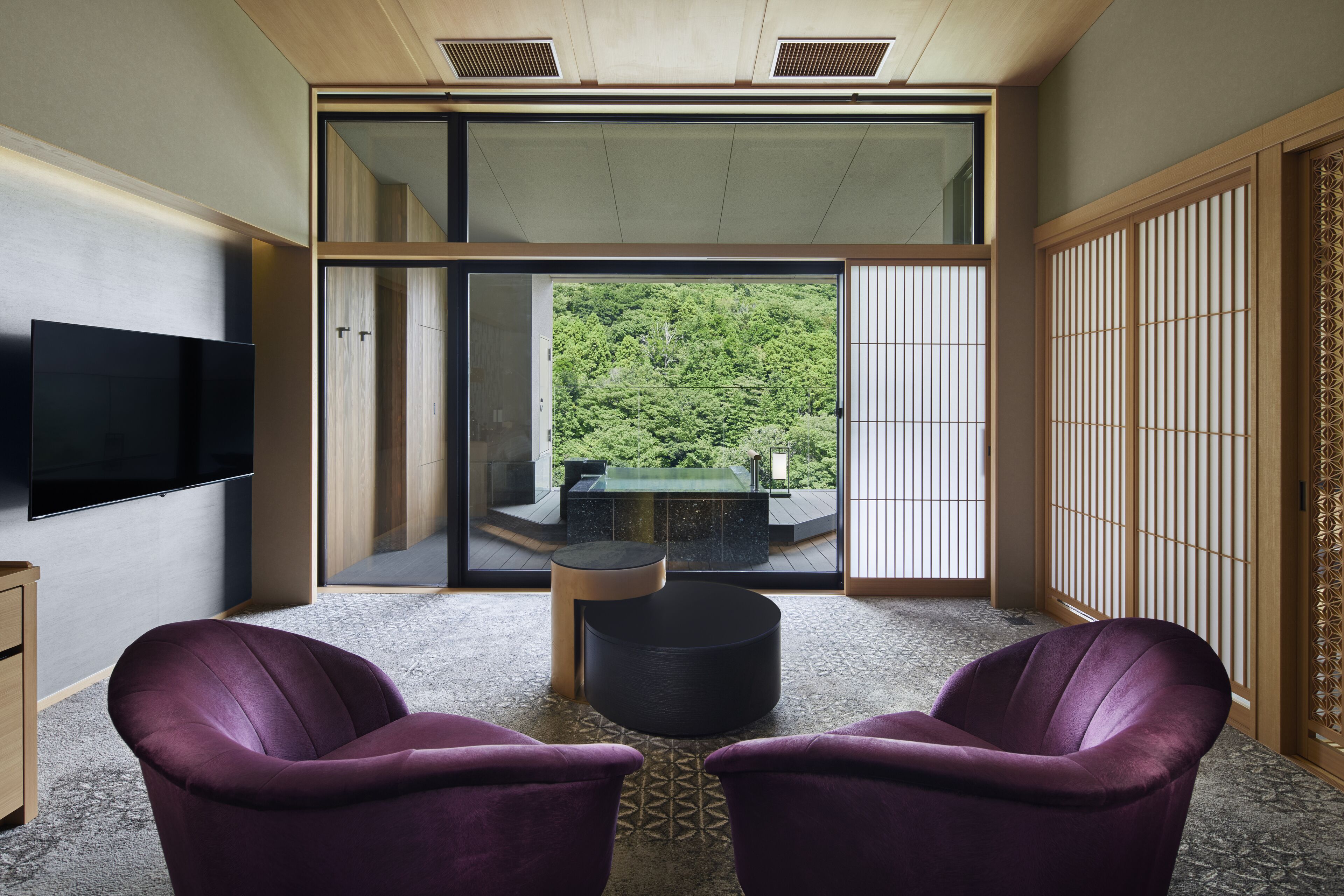 A room with a large sliding glass door leading to a balcony with a view of a forest. There are two purple chairs and a small round table in the room.