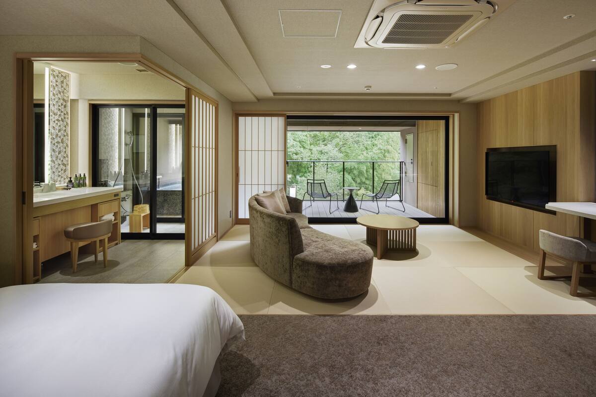 A modern hotel room with a large bed, a sofa, a desk with a chair, a TV, and a balcony with a view of greenery.