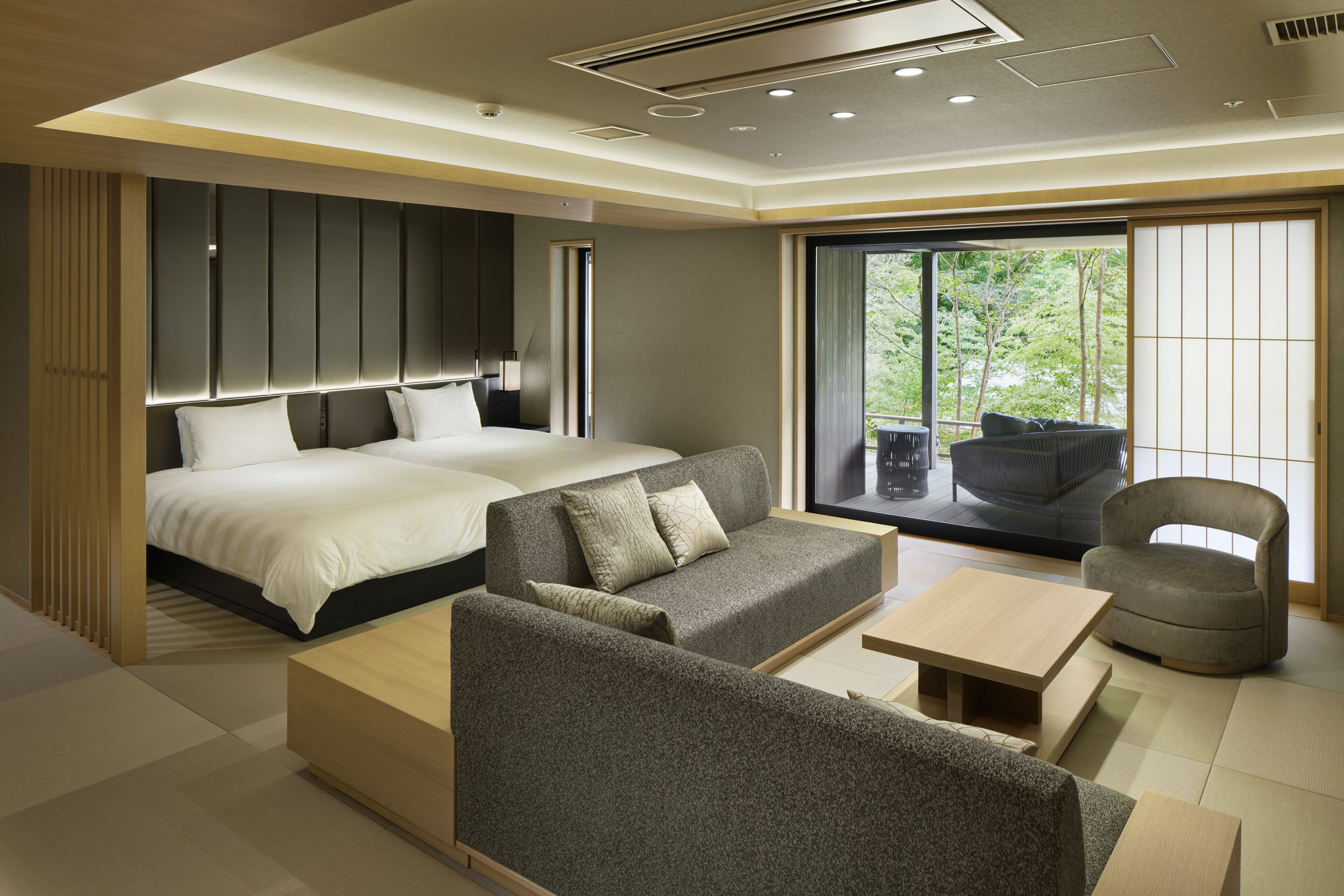 A modern hotel room with a large bed, a sofa, a small table, and a chair. There is a sliding door leading to a balcony with a view of trees.
