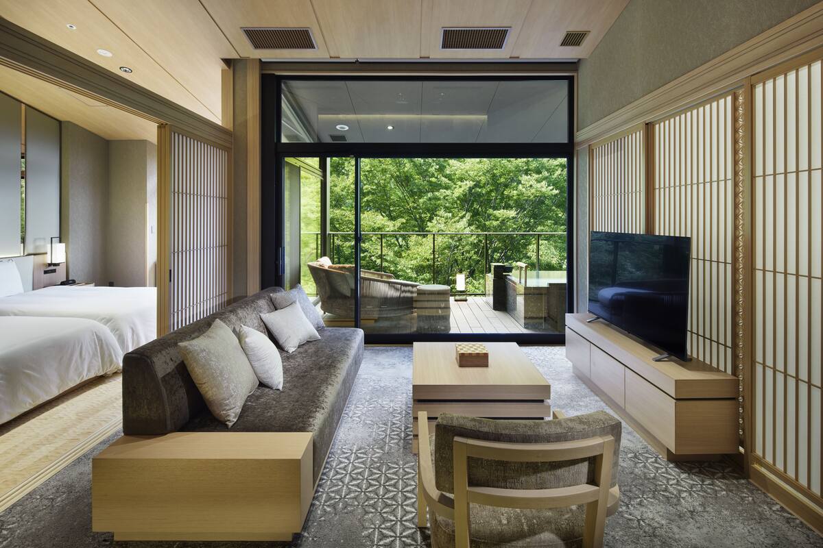 A modern hotel room with a sofa, a TV, a dining table, and a balcony with a view of trees.