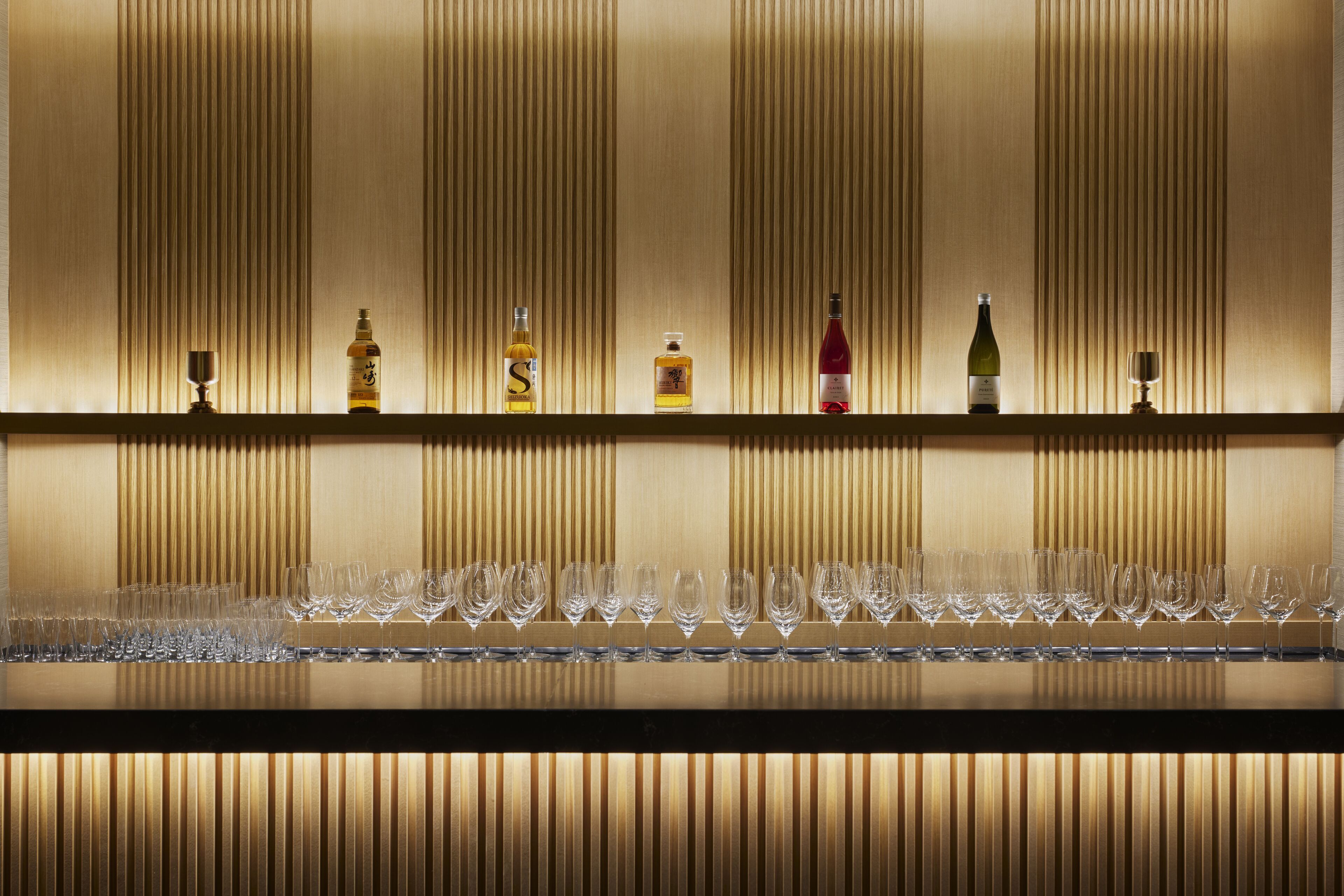 A bar area with a row of wine glasses and bottles of liquor on a shelf against a wall with vertical lines.