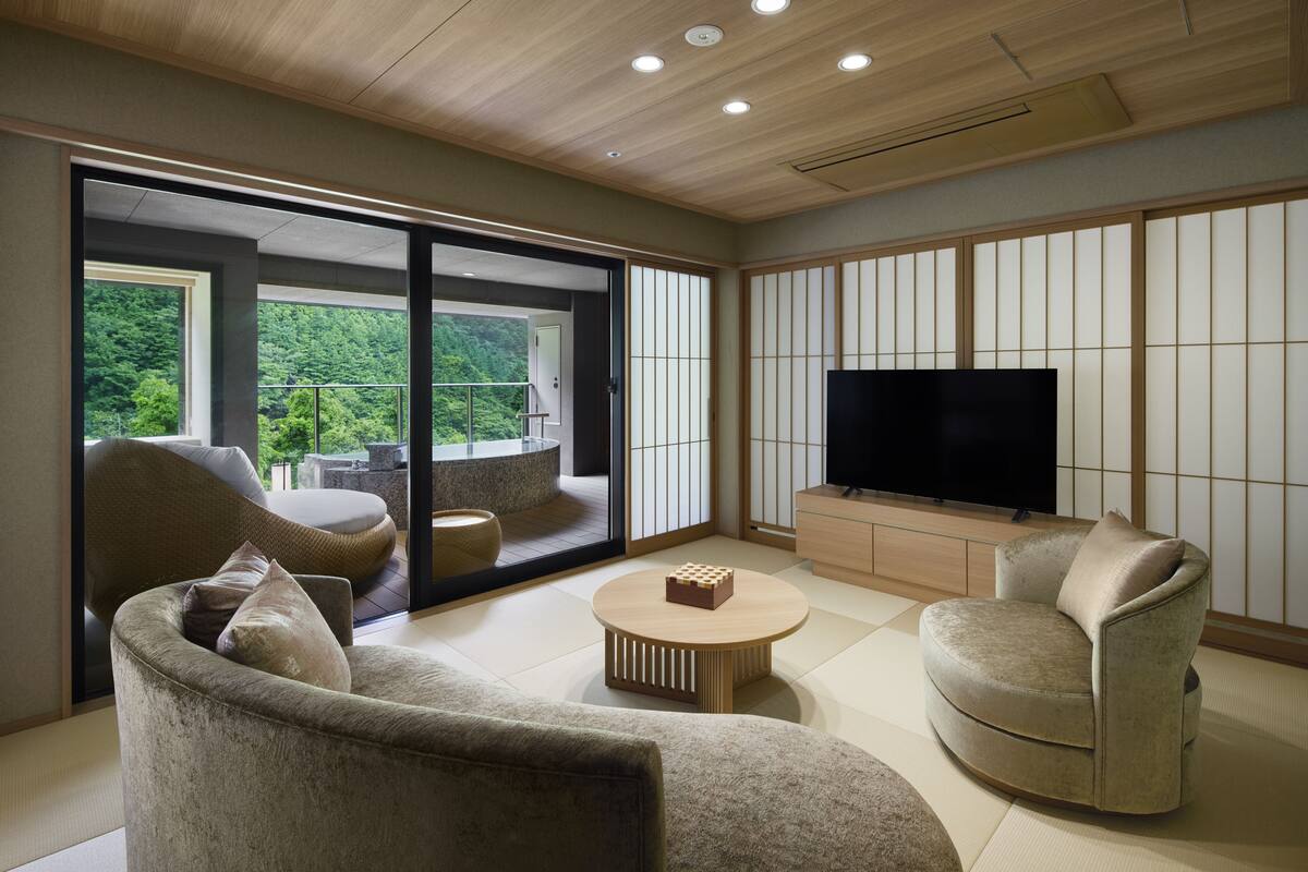 A modern living room with a large TV, comfortable seating, and a view of greenery outside.