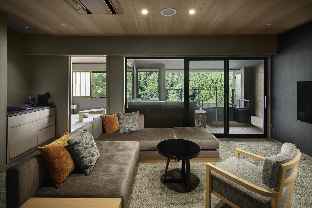 A modern living room with a sectional sofa, a round coffee table, and a wooden chair. There is a large sliding glass door leading to a balcony with a view of greenery.