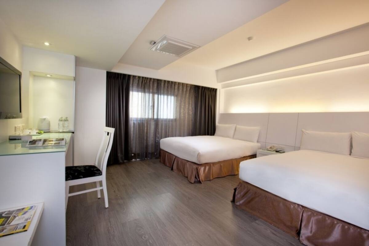 A hotel room with two beds, a desk with a chair, a large window with curtains, and a modern ceiling design.
