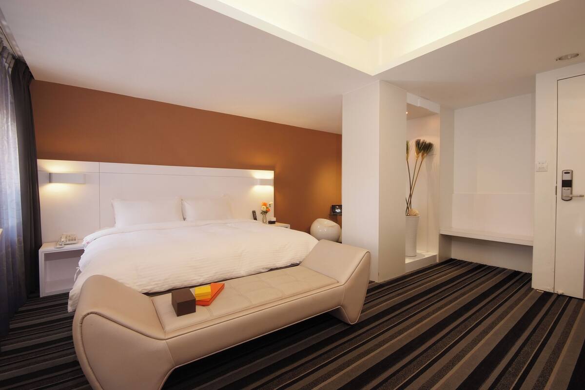 A modern hotel room with a large bed, a bedside table with a lamp, a small seating area, and a wardrobe.