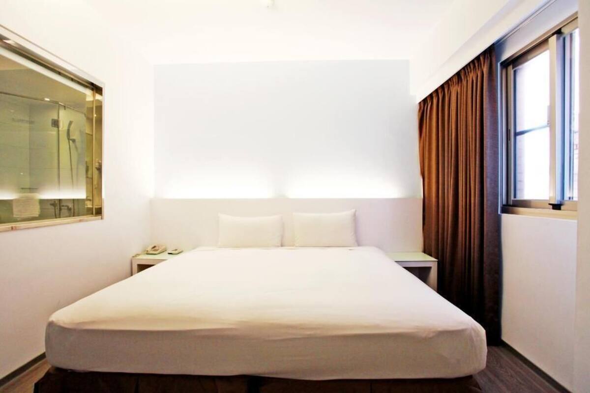 A hotel room with a large bed, a bedside table with a lamp, a mirror, and a window with curtains.