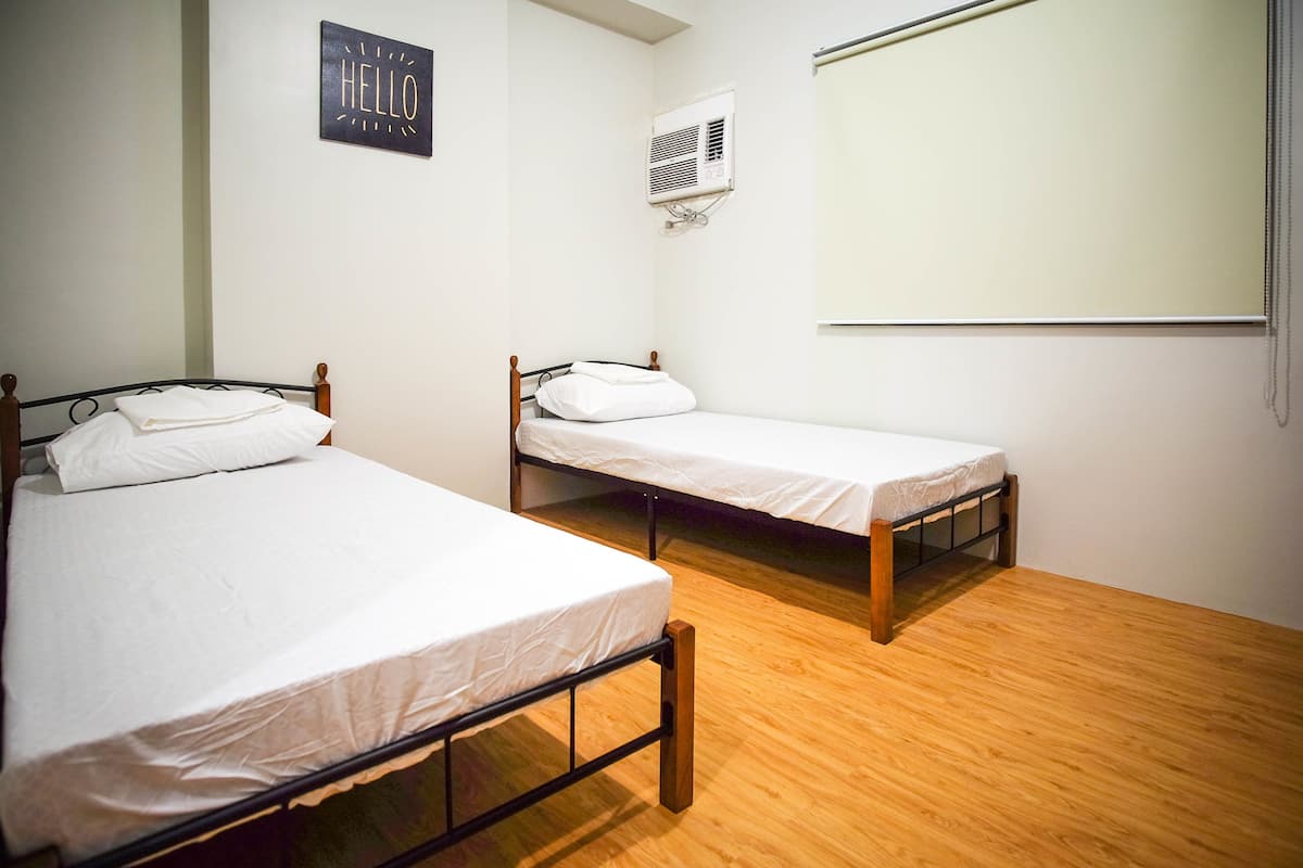 Duas camas de solteiro com estruturas de madeira e roupa de cama branca, um ar condicionado fixado na parede e um letreiro com a palavra 'HELLO'.