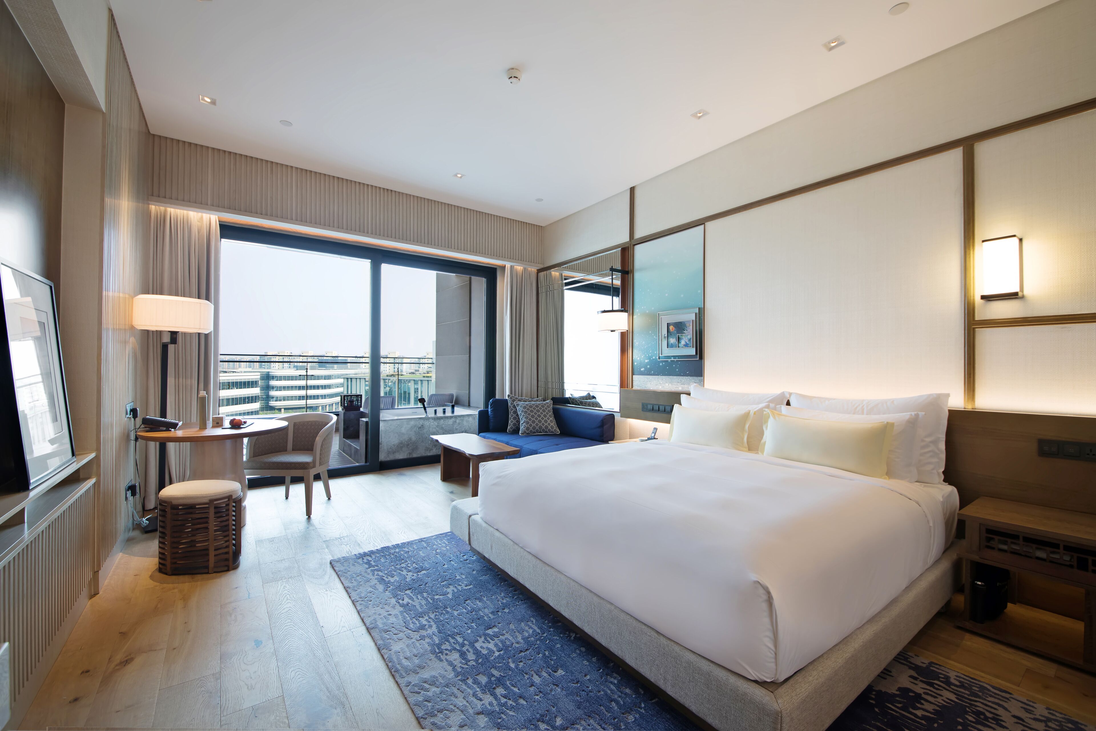 A modern hotel room with a large bed, a sitting area with a sofa and armchair, a small table, and a view of the city through the window.