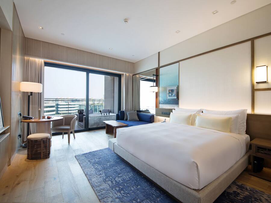 A modern hotel room with a large bed, a sitting area with a sofa and armchair, a small table, and a view of the city through the window.