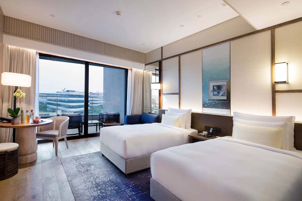 A hotel room with two beds, a desk with a chair, a small table with a plant, and a large window with a city view.
