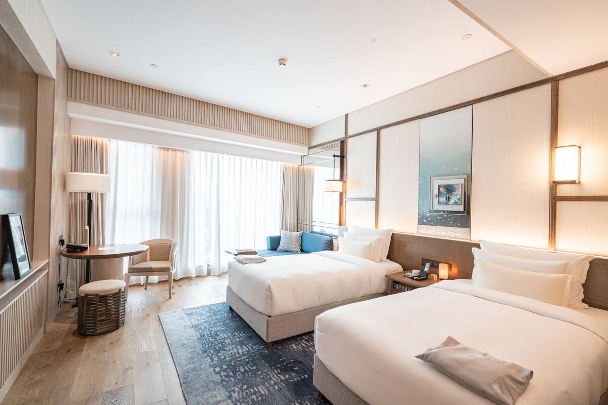 A hotel room with two beds, a sitting area with a table and chairs, a large window with curtains, and a wall-mounted TV.
