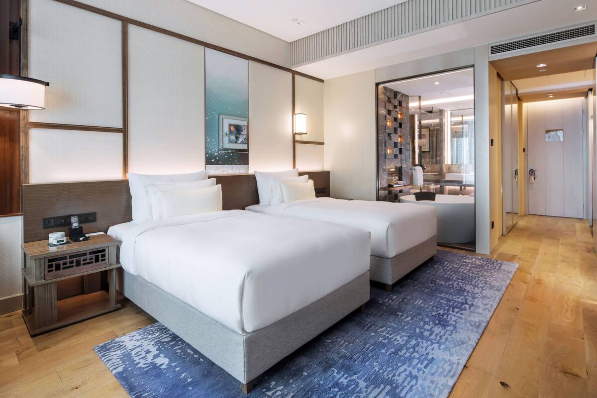 A hotel room with two beds, a nightstand, a mirror, and a bathroom visible through an open door.