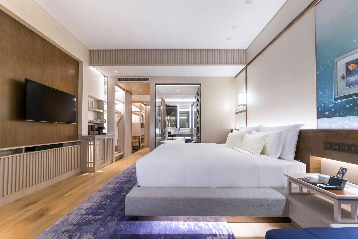 A modern hotel room with a large bed, a flat-screen TV, a desk with a laptop, and a view of the hallway.
