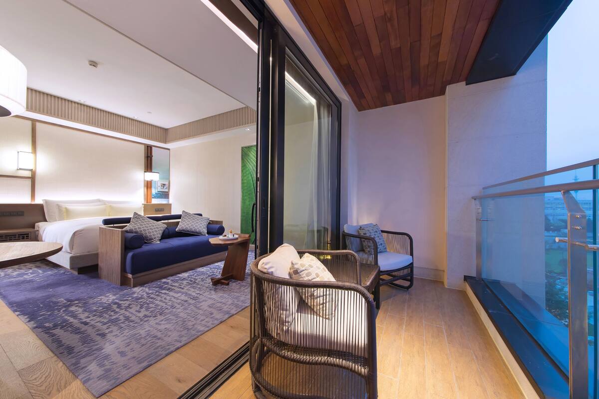 A modern hotel room with a balcony, a bed, a sofa, and a wicker chair.