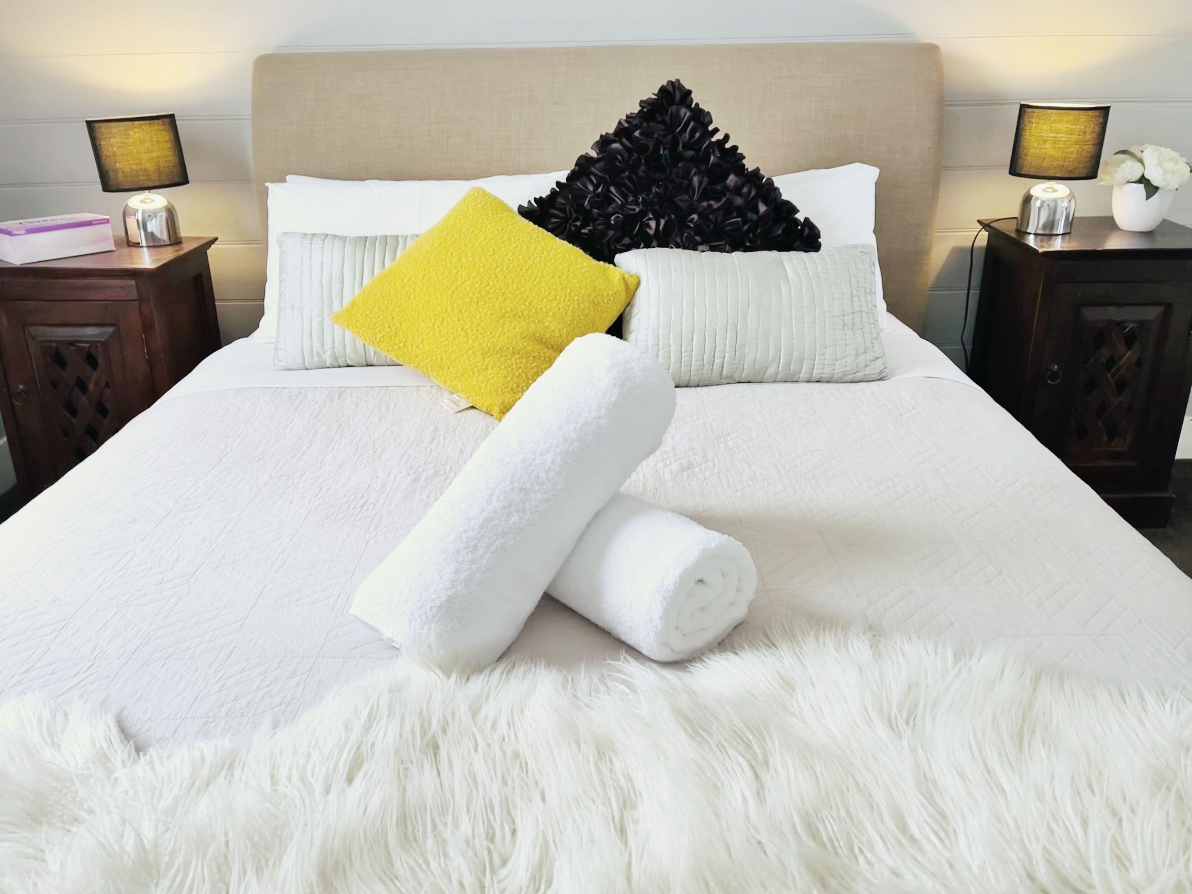 A neatly made bed with white and yellow pillows, a fluffy white rug, and two bedside tables with lamps.