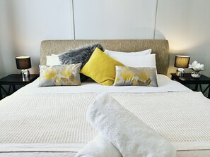 Una cama bien hecha con un edredón blanco, varios almohadones en amarillo y blanco, y un plaid gris mullido.