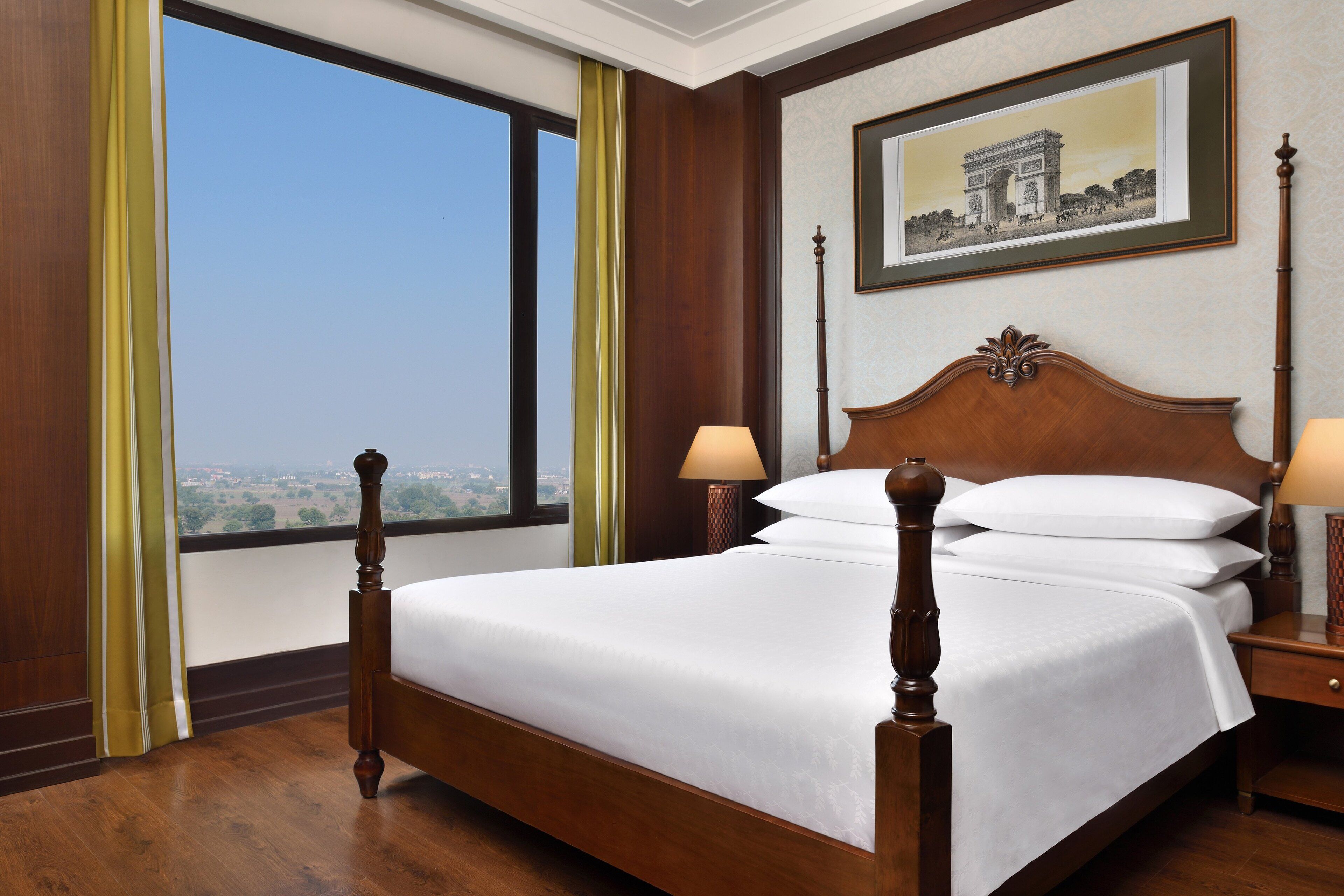A neatly made bed with a wooden headboard, two bedside lamps, and a framed picture of the Arc de Triomphe on the wall.