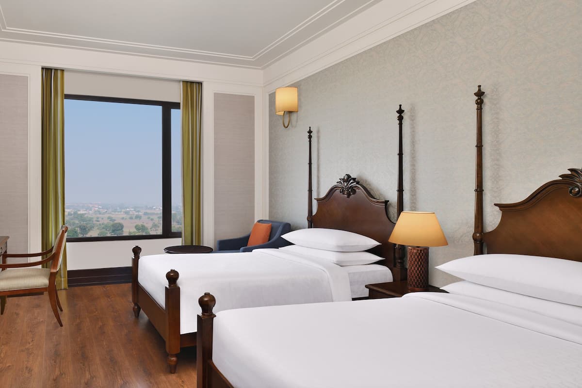 A hotel room with two beds, a chair, a nightstand with a lamp, and a view of the city through a large window.