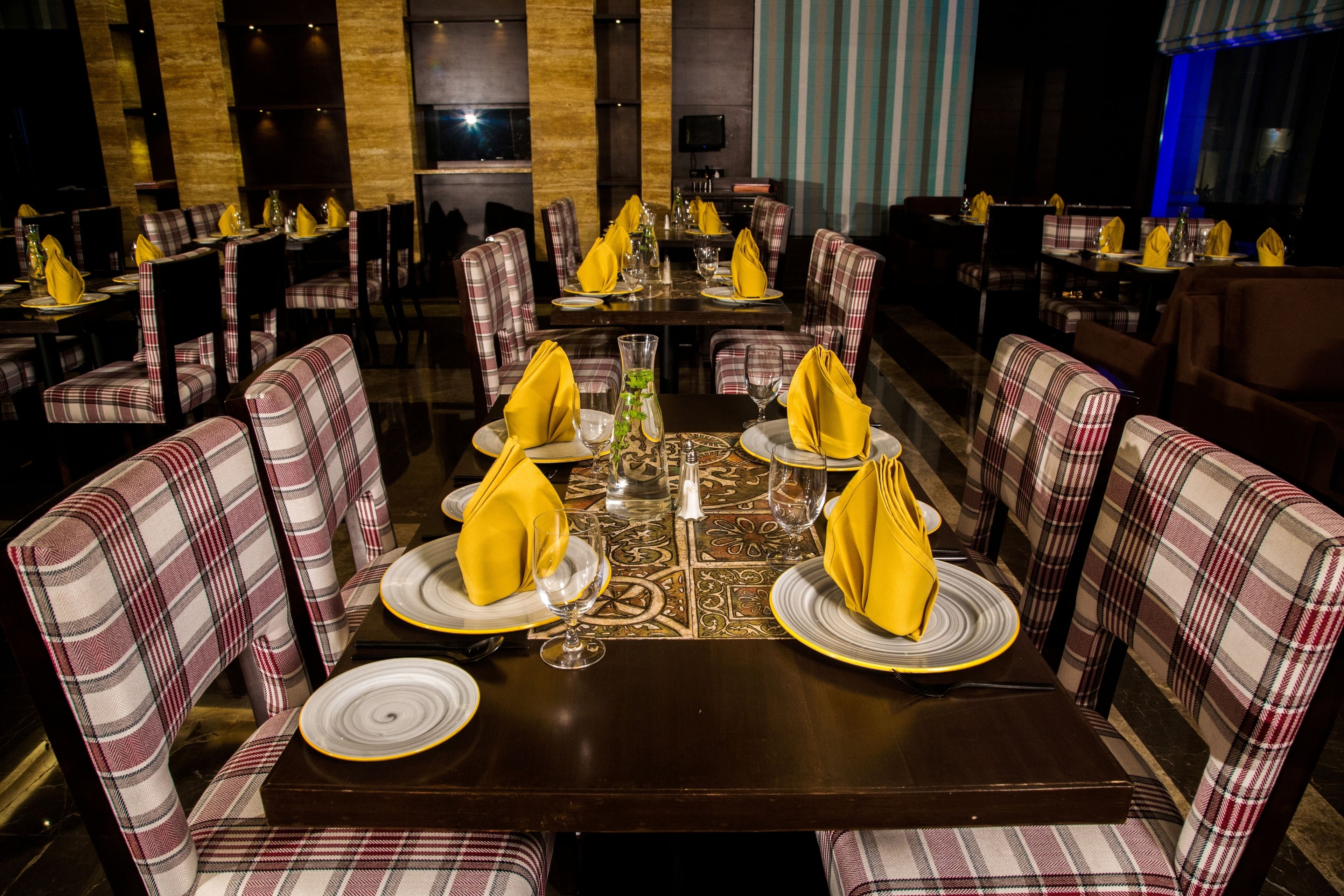 A restaurant with multiple tables set with yellow napkins, white plates, and clear glasses. The chairs have a plaid pattern.