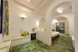 Hotelkamer met een vloer van groene tegels, een bed met een bruine dekbedovertrek, een klein bureau met een lamp, een bankje met kussens, en een douchegedeelte met een glazen deur.