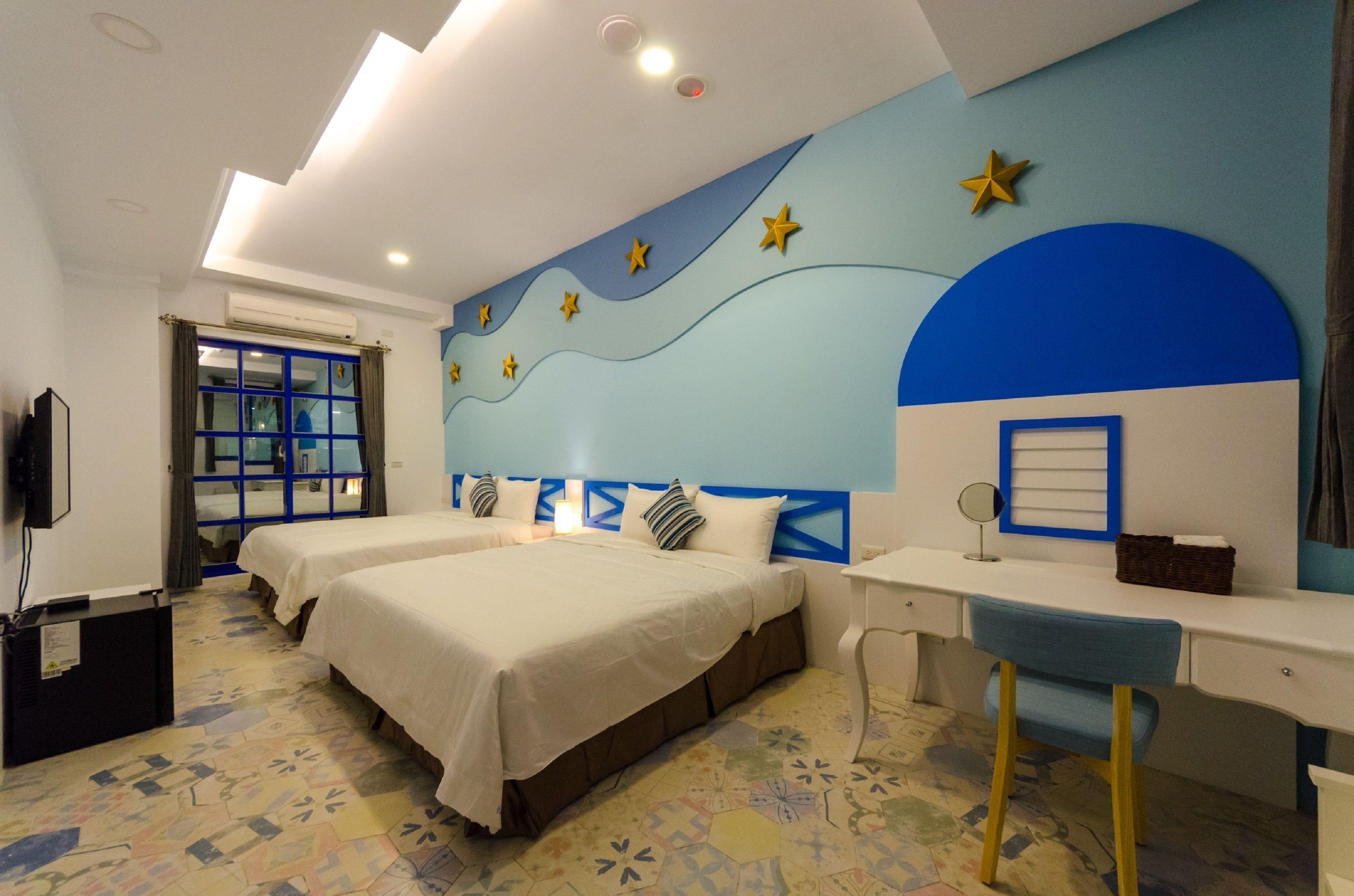 A bedroom with two beds, a desk, and a chair. There is a window with blue curtains and a wall with star decorations.