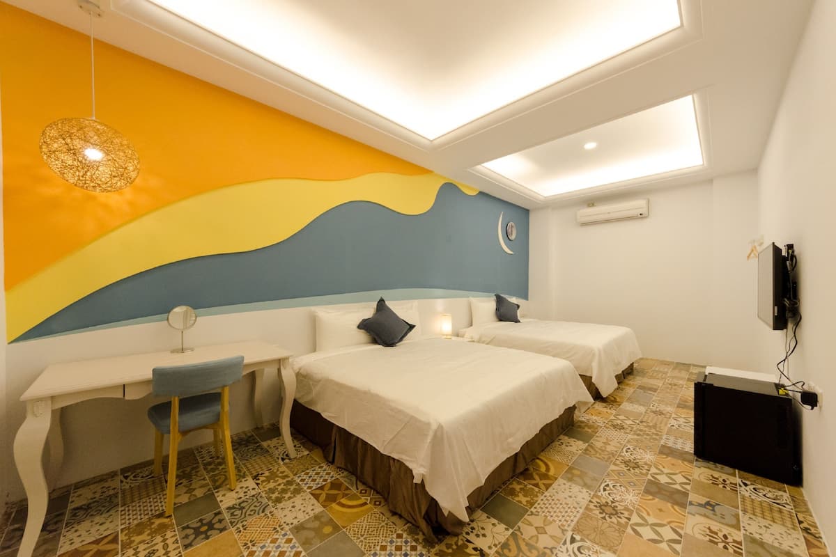 A hotel room with two beds, a desk, and a chair. There is a colorful mural on the wall, a ceiling light, and a flat-screen TV mounted on the wall.