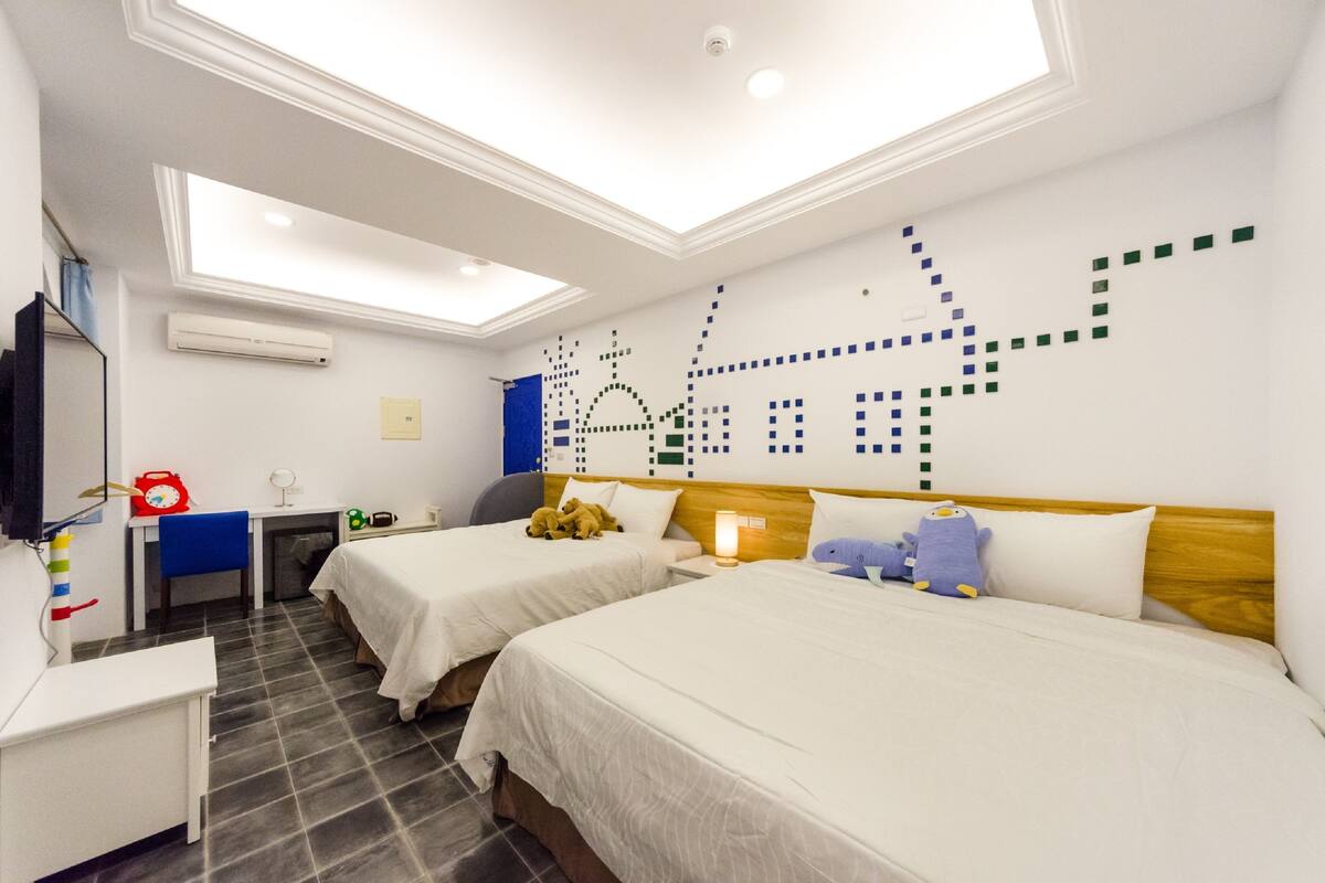 A hotel room with two beds, a desk, a chair, a TV, and a ceiling with a geometric light fixture.