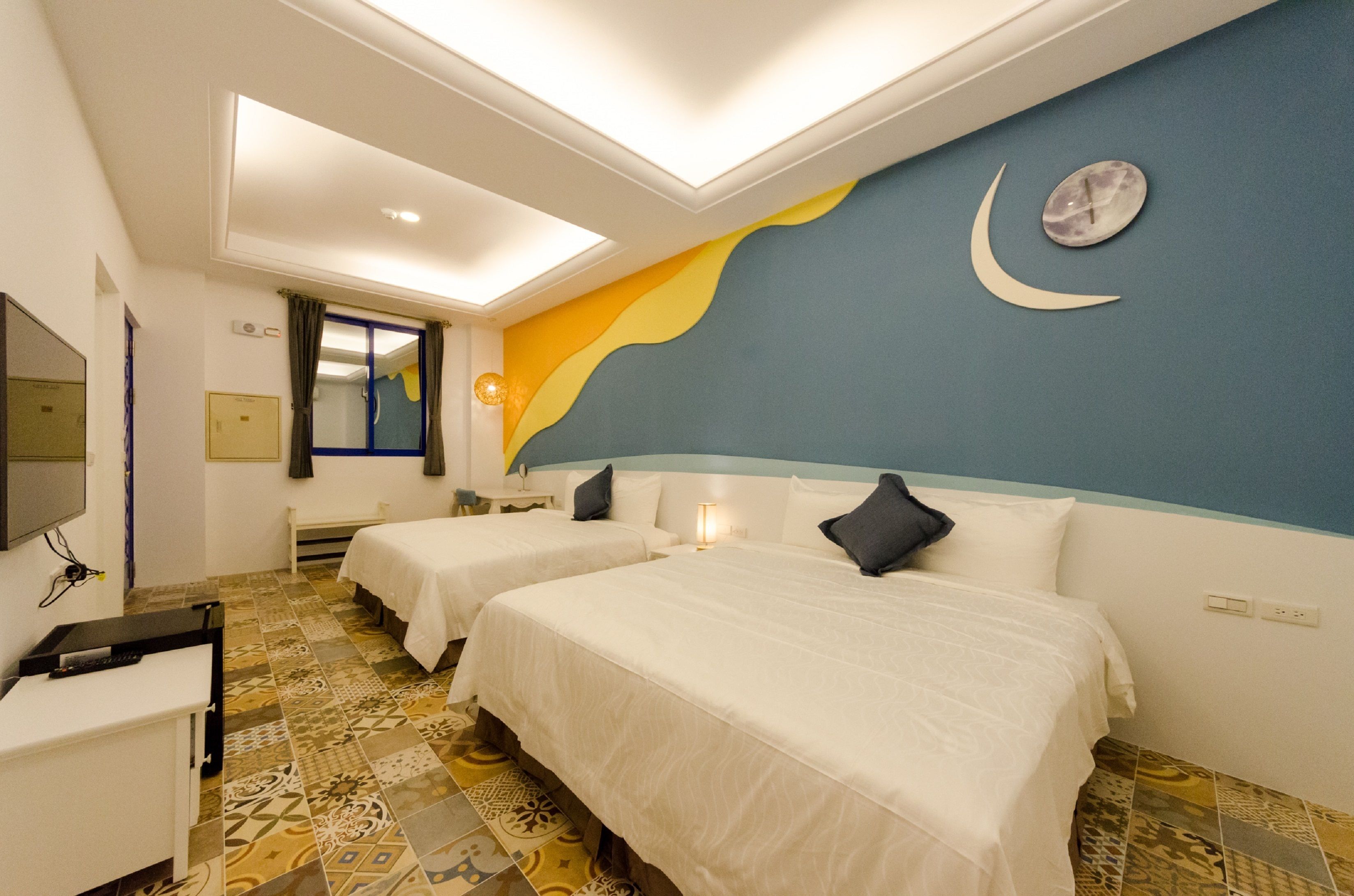 A hotel room with two beds, a desk, a TV, and a wall with a crescent moon and abstract art.