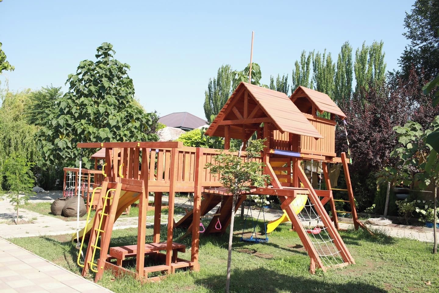 A wooden play structure with multiple levels and slides, surrounded by greenery and trees.