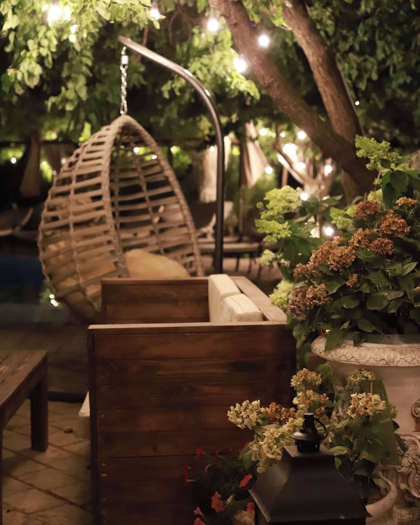 Zona d'estar a l'aire lliure amb bancs de fusta i una cadira penjant, envoltada de vegetació i il·luminació decorativa.
