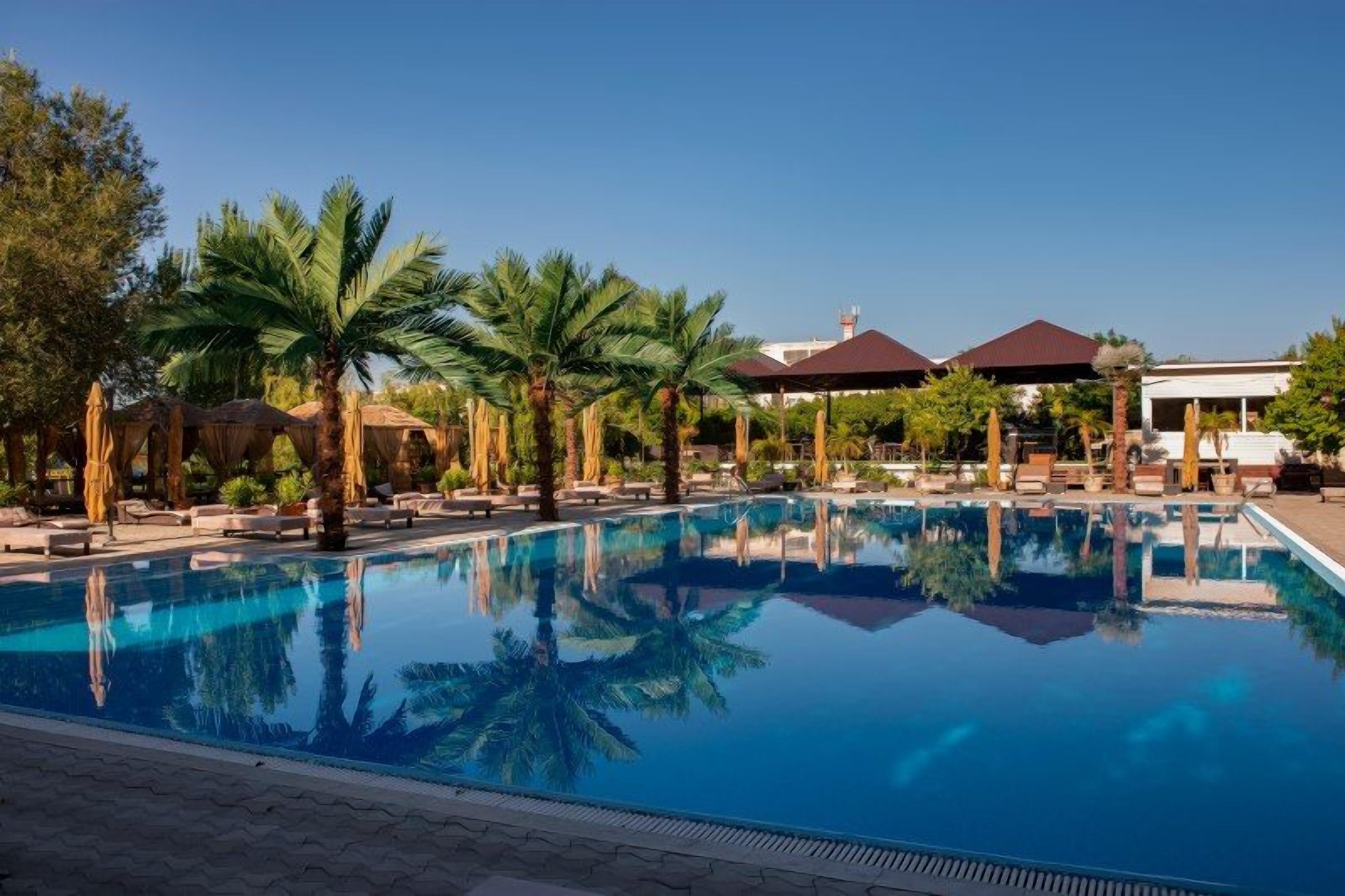 Une piscine aux eaux cristallines, bordée de chaises longues et de palmiers, le tout entouré de bâtiments aux toits rouges.