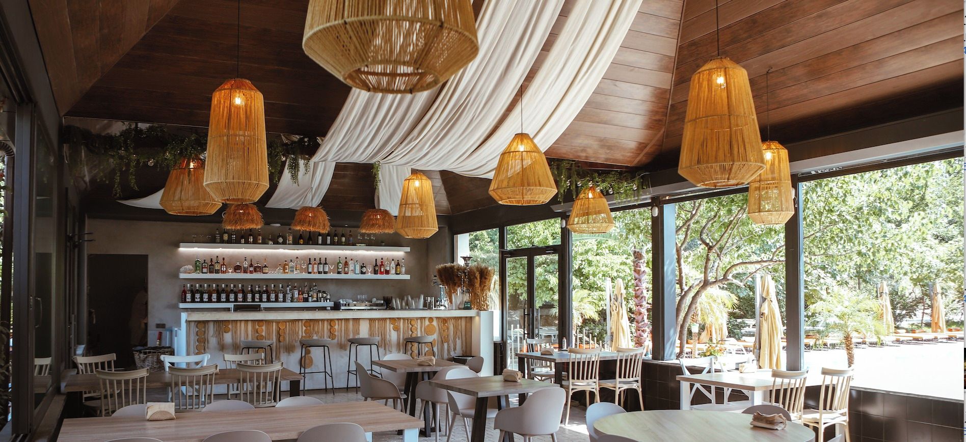 Interior d'un restaurant amb lluminàries de vimet penjades, una zona de bar amb estanteries plenes de botelles i taules i cadires disposades per dinar.