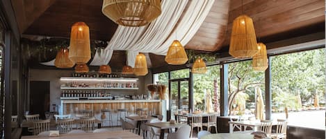 A restaurant interior with hanging wicker lamps, a bar area with shelves of bottles, and tables and chairs arranged for dining.