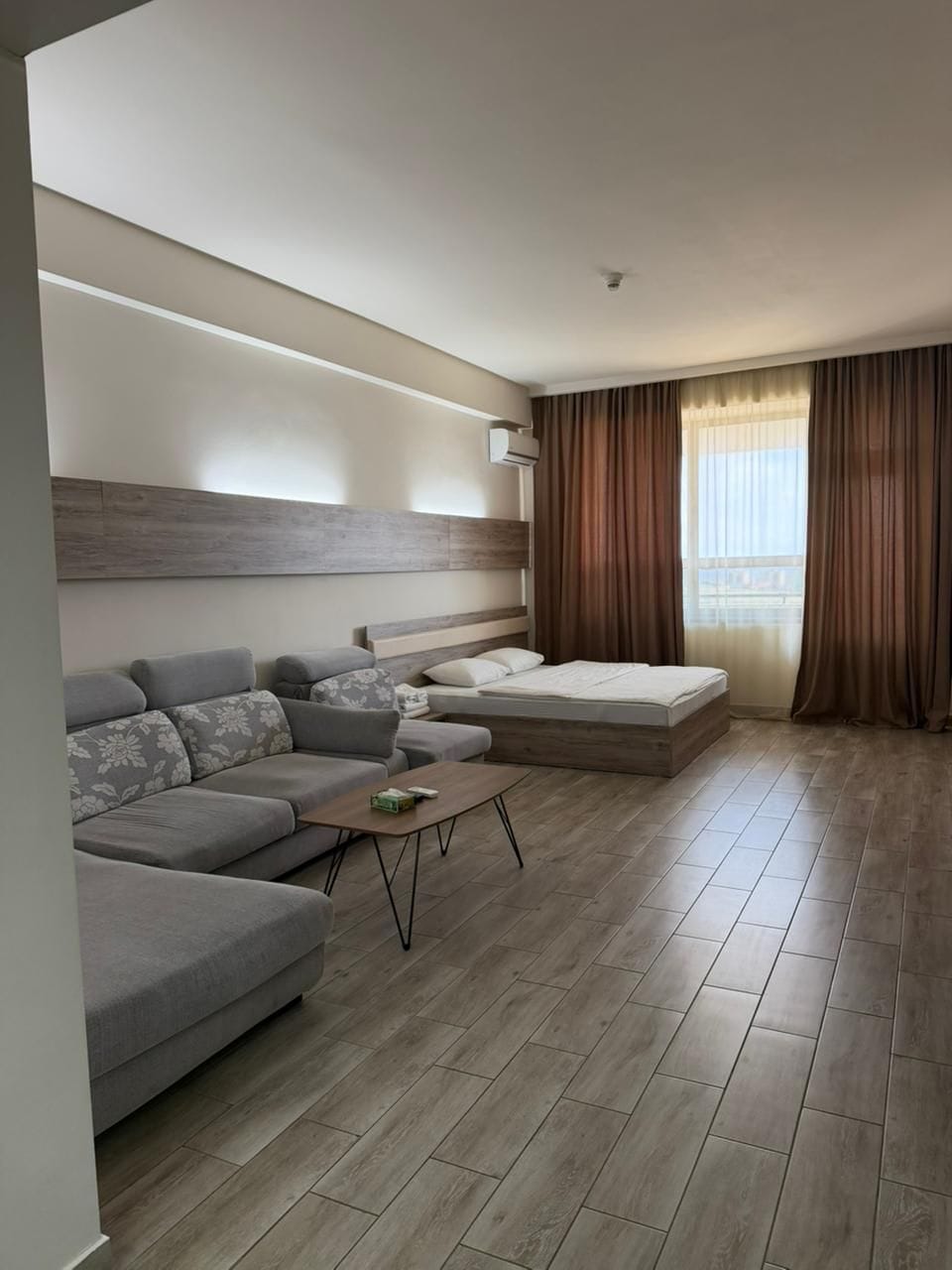 Een moderne hotelkamer met een bank, een bed, een bijzettafeltje en een groot raam met gordijnen.