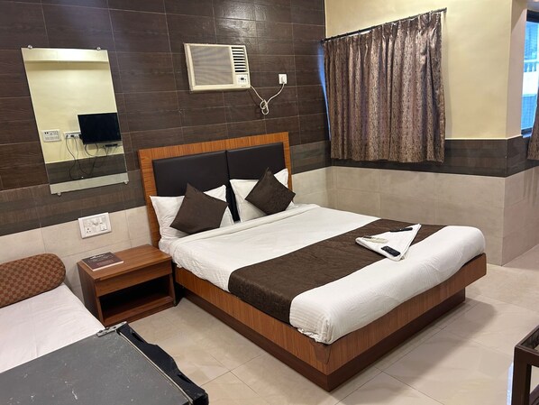 A hotel room with a large bed, a bedside table, a mirror, an air conditioning unit, and a window with curtains. - Hotel Lucky Bandra (Mumbai)