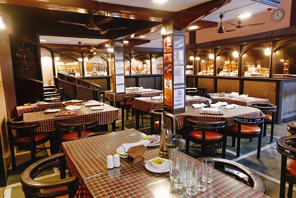 A restaurant interior with multiple tables and chairs, a staircase, and a wall displaying various items. - Hotel Lucky Bandra (Mumbai)
