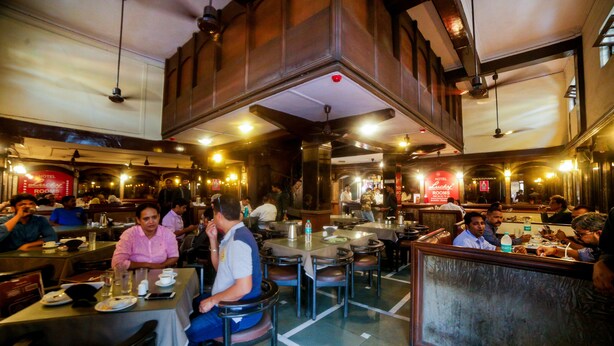 Een bruisend restaurant met meerdere eettafels en stoelen, een balie en een hoge plafond met zichtbare balken.