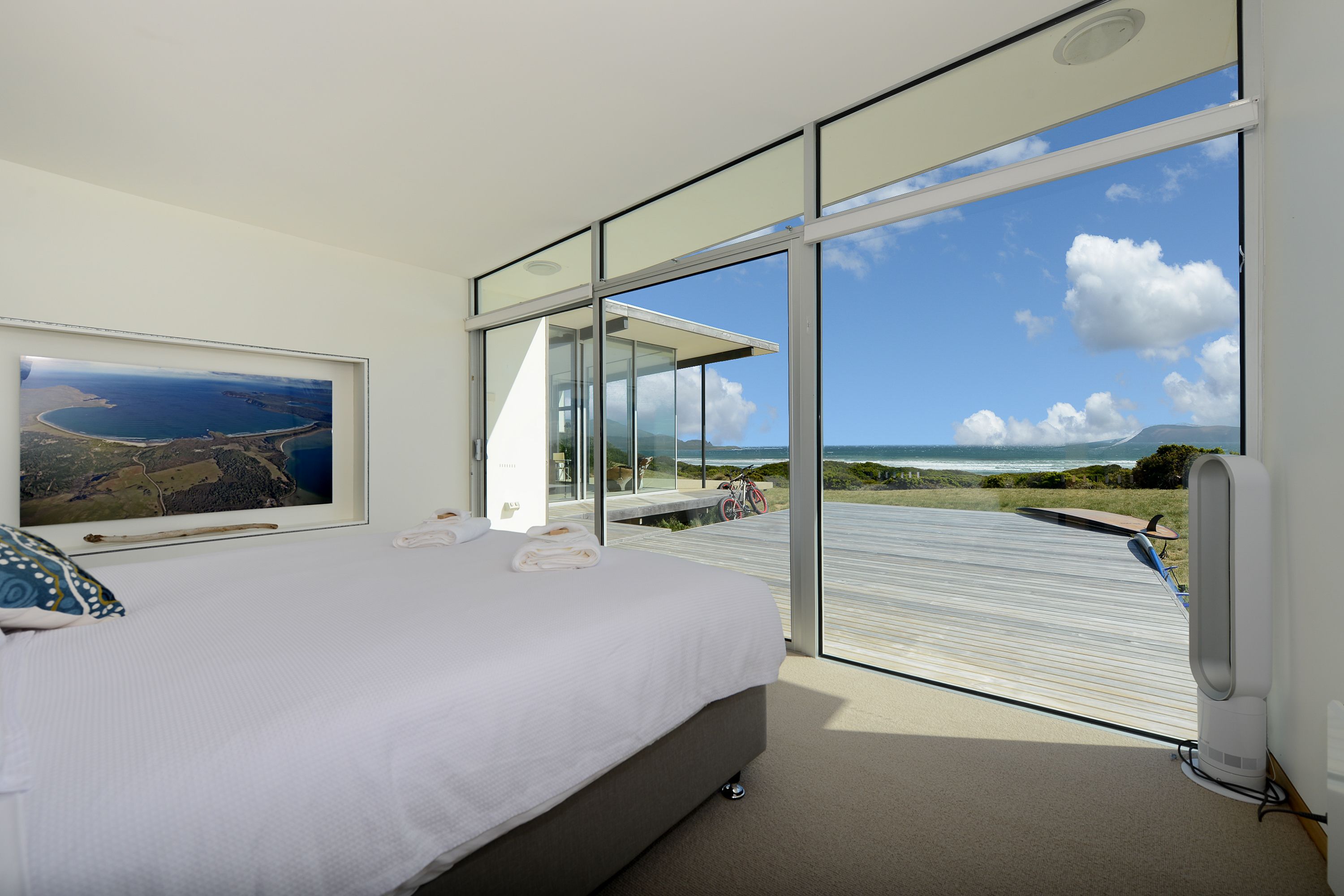 A bedroom with a large bed, a framed landscape photo, and a view of the ocean through floor-to-ceiling windows.