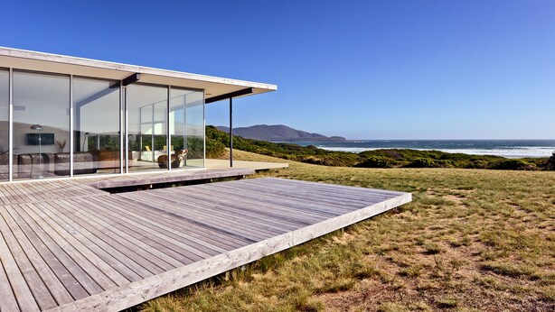 A modern house with a wooden deck and large glass windows overlooking the ocean.